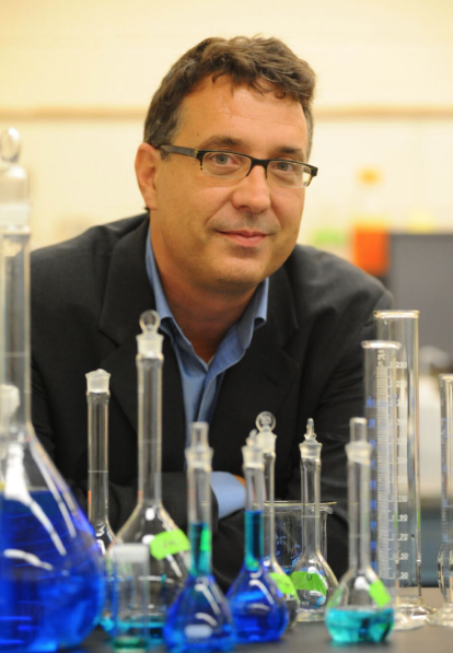 A man, smiling and wearing glasses, sits a a table filled with beakers holding brightly colored liquids