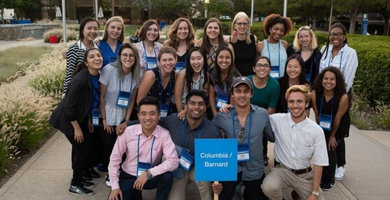 A group of roughly two dozen people poses on a sidewalk with a placard reading "Columbia/Barnard"