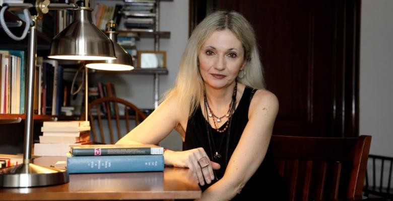 A woman sites at a writing desk the top of which is covered with books and flanked by two reading lamps