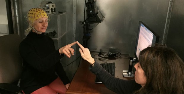 Two women face each other with index fingers touching, as one wears a headpiece of electrical wires attached to computer equipment