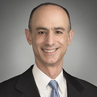 A head shot of a man wearing a dark suit and pale blue tie smiling into the camera