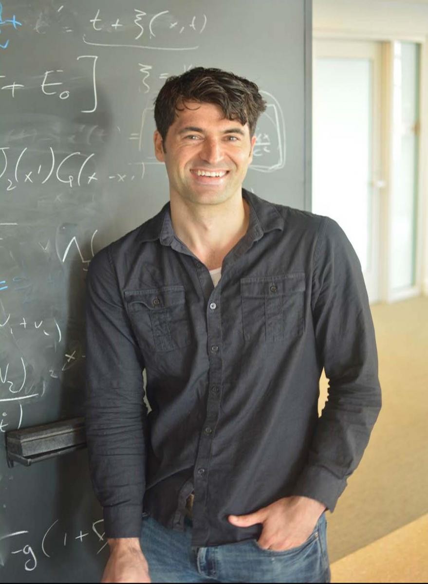 Wearing a black button-downed shirt and jeans, a man stands against a blackboard, smiling, hand in pocket