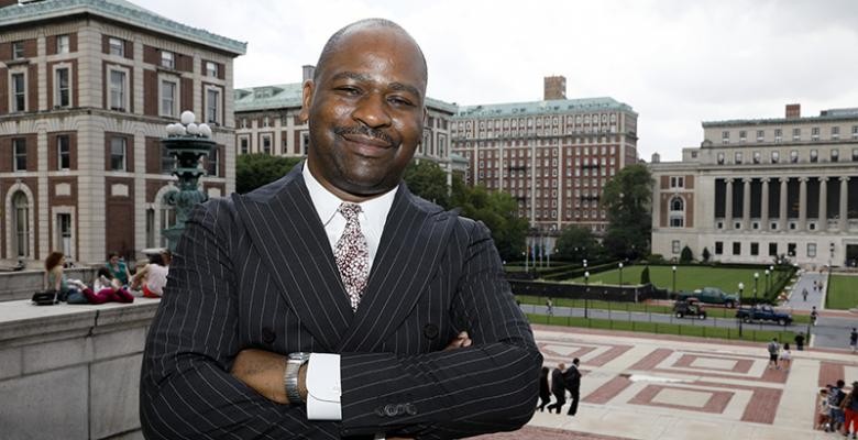 A man stands on the steps of Low Library wearing a dark suit and with his arms folded across his chest; he smiles into the camera