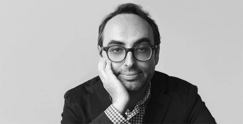 A man, wearing glasses and looking directly into the camera, rests his head against the palm of his right hand