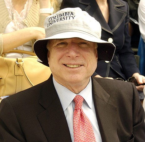 John McCain wears a dark suit and red print tie with a bucket hat printed with "Columbia University" to shield against the bright sun.