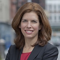 A head shot of a woman standing on a New York City street, wearing a dark gray suit coat, and smiling into the camera