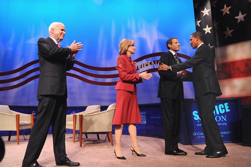 On a stage, John McCain and two other people greet President Obama as he walks toward them.
