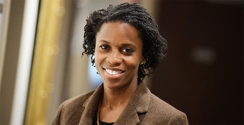 Wearing a brown, wool blazer, a woman, with her hair pulled back into a loose ponytail, smiles into the camera
