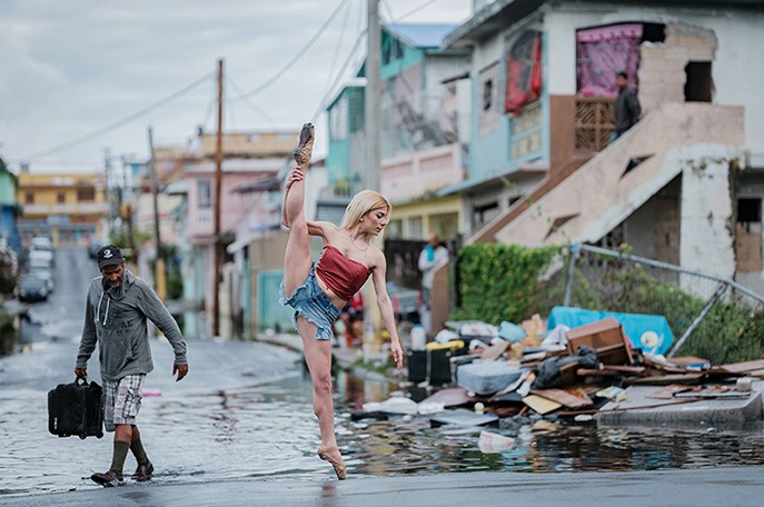Standing in the middle of a flooded street, a ballerina dressed in blue jean shorts and a bandeau top stands on pointe on her left leg and lifts her right into a 180-degree split.