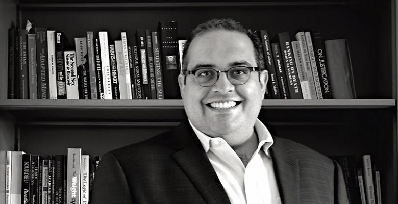 Wearing a blazer and dress shirt, a man stands in front of shelves lined with books, smiling