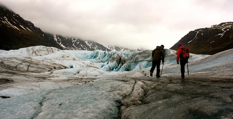 Two people trek across the surface of a glacier, high in the clouds