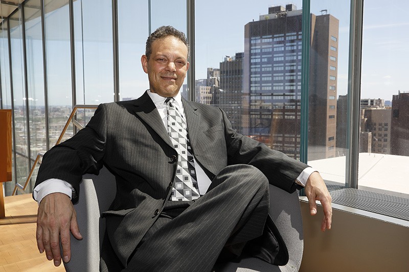 Dressed in a suit and tie, a man relaxes in an armchair with a wall of windows showing the skyline of Upper Manhattan behind him.