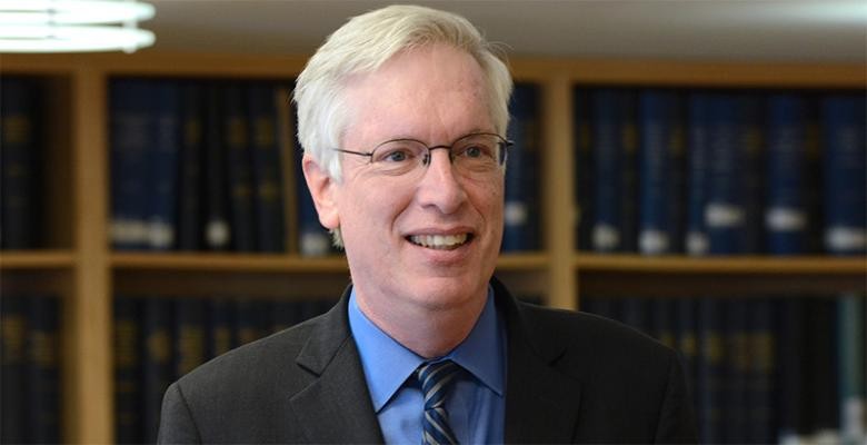 Wearing a dark suit and tie, a man stands in front of shelves lined with books, smiling into the camera