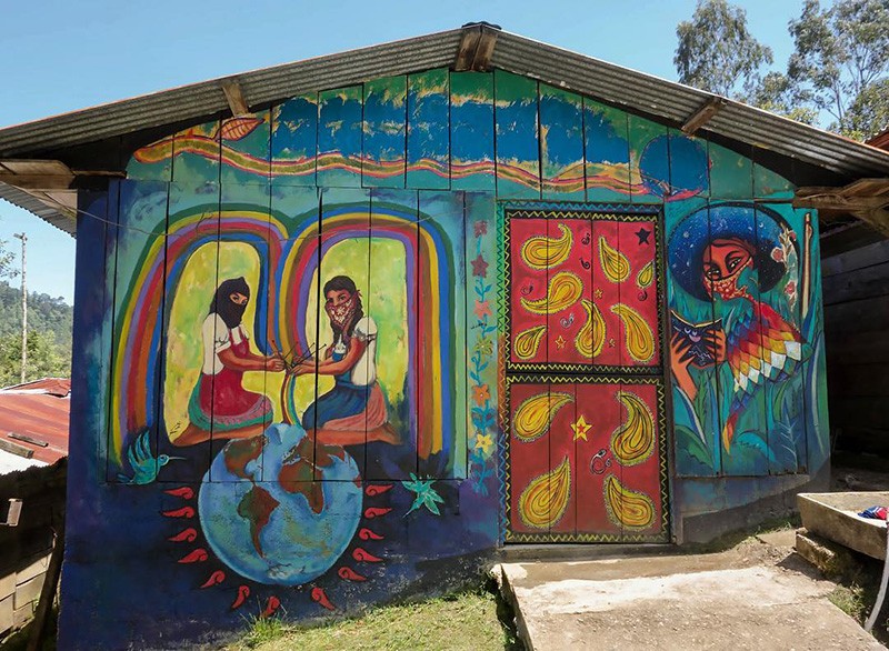 The facade of a small building is covered by a colorful mural depicting two women, kneeling and facing each other. The women stretch their hands toward each other.