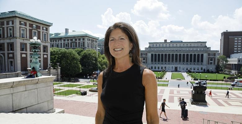 Wearing a sleeveless, black dress, a woman stands on the steps of Low Library, smiling into the camera on a bright sunny day