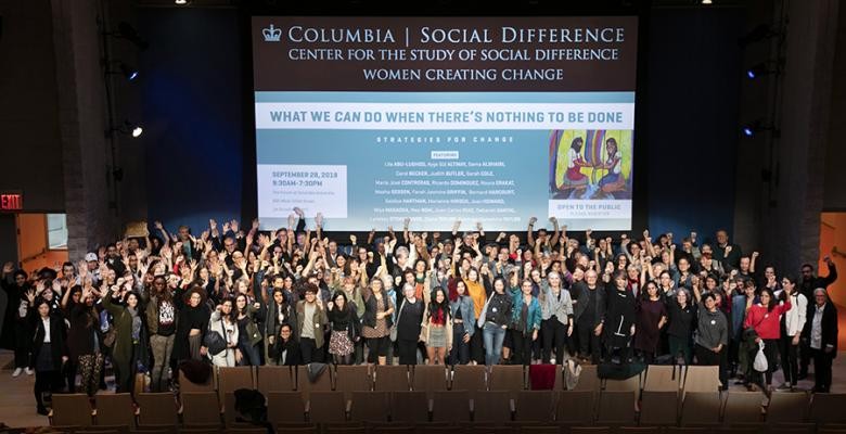 A group of nearly 100 women, many with an arm raised in defiance, stand on stage beneath a banner that asks "What can we do when there's nothing to be done?"