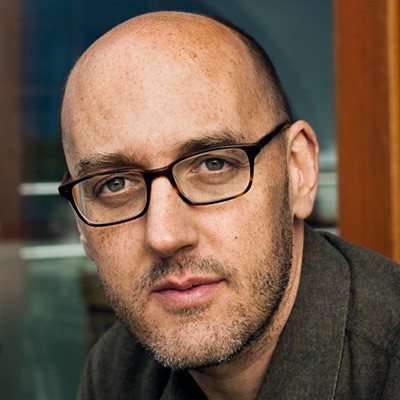 A closeup head shot of a man wearing glasses and looking directly into the camera pensively