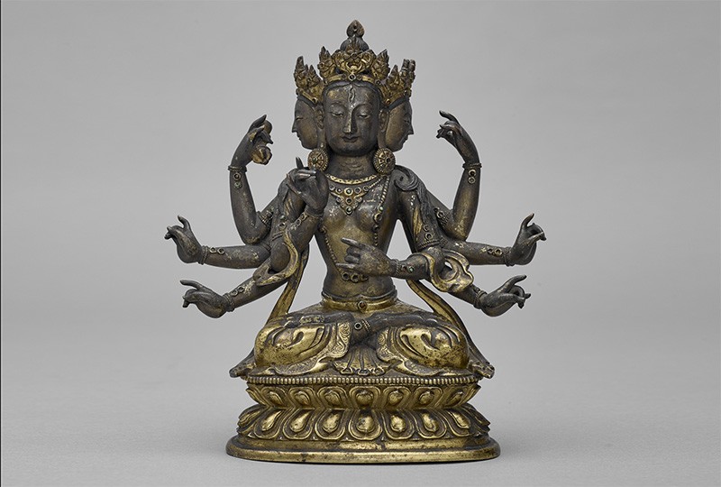 A bronze statue of of goddess with six arms and an elaborate headdress, seated on a base