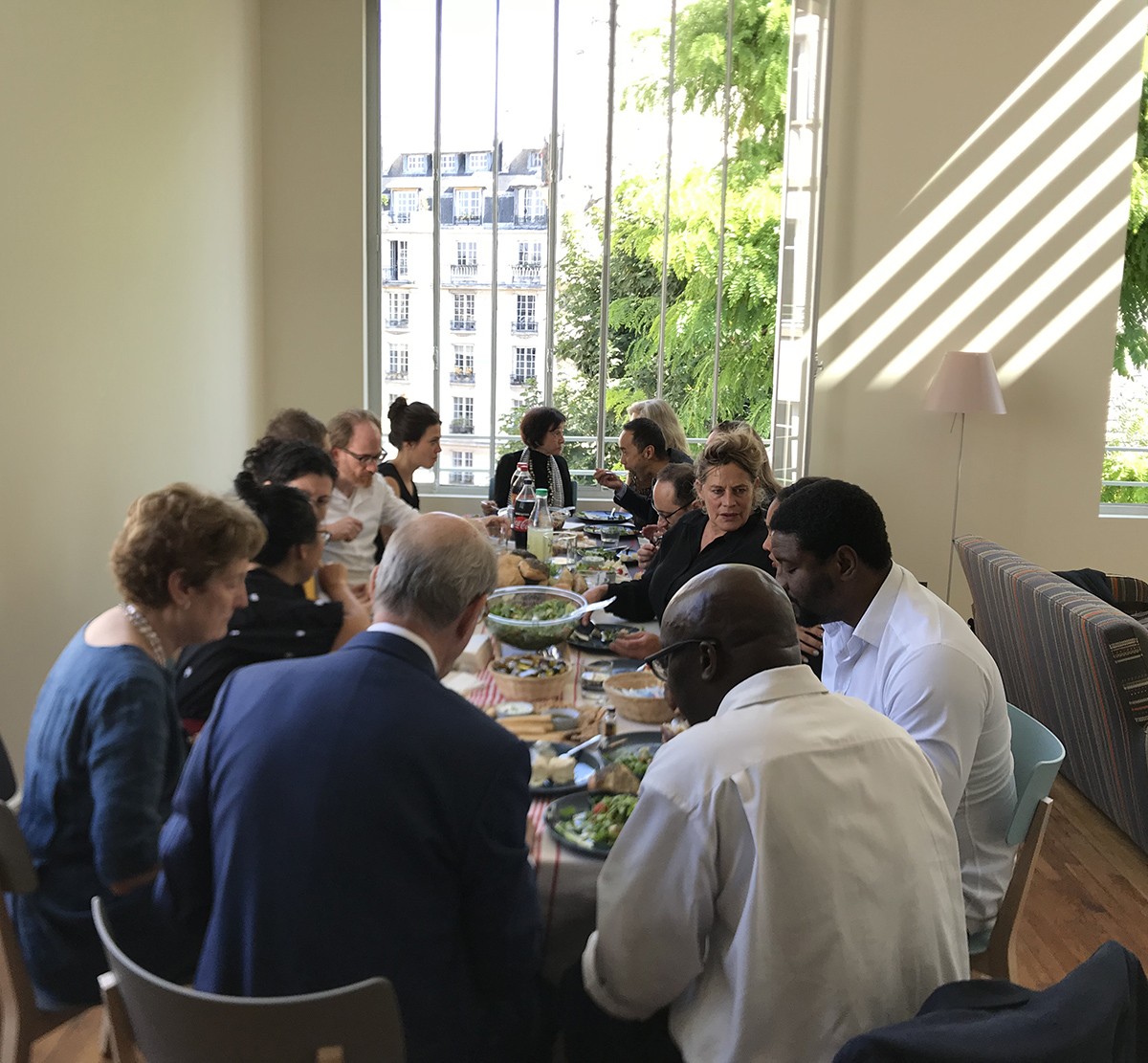 Photo graph of a group of people sitting down to lunch inside a big room, in front of a big window with light streaming in