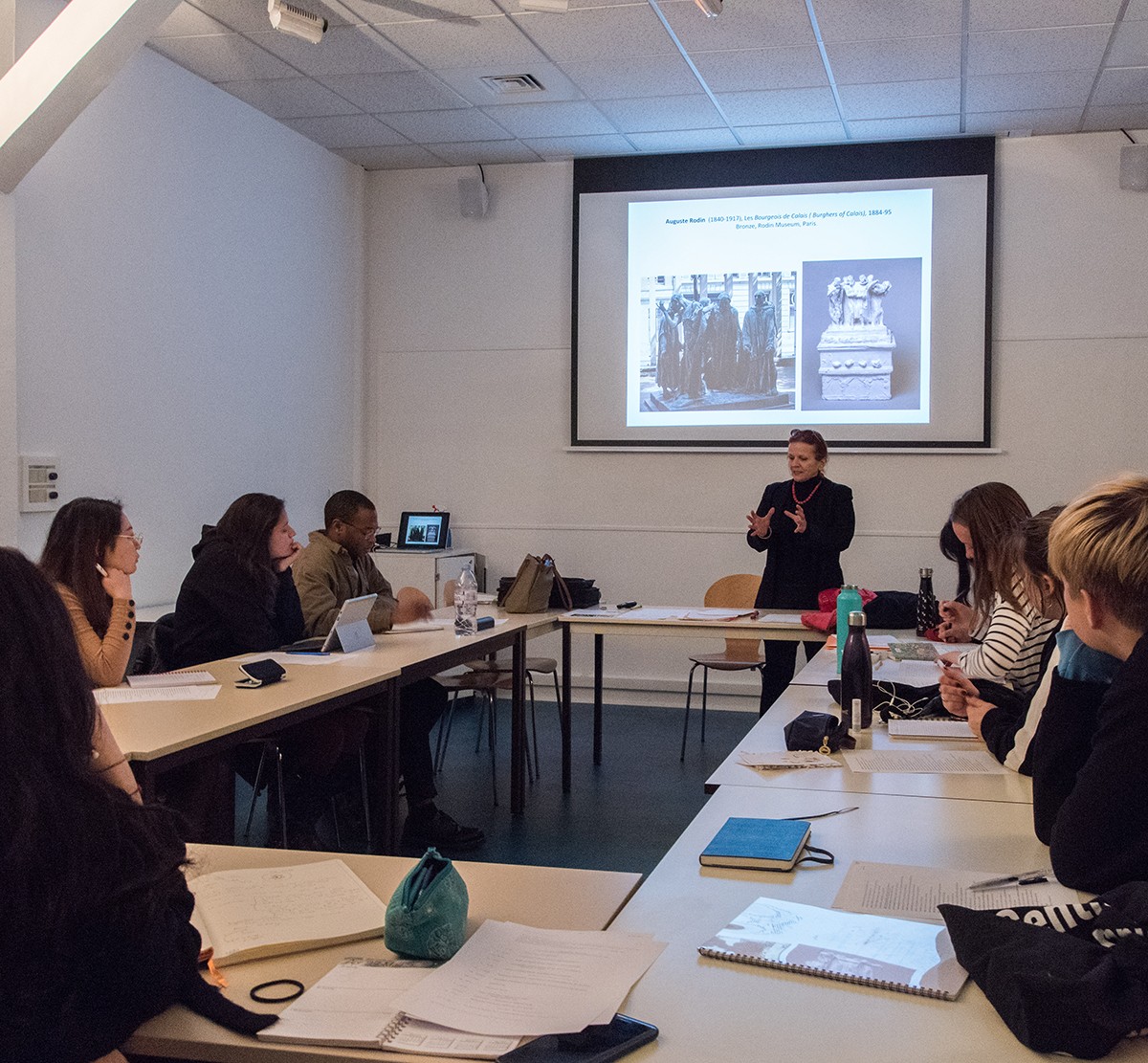 Photo of a classroom with desk in a horseshoe, filled with students with a screen projector in the background