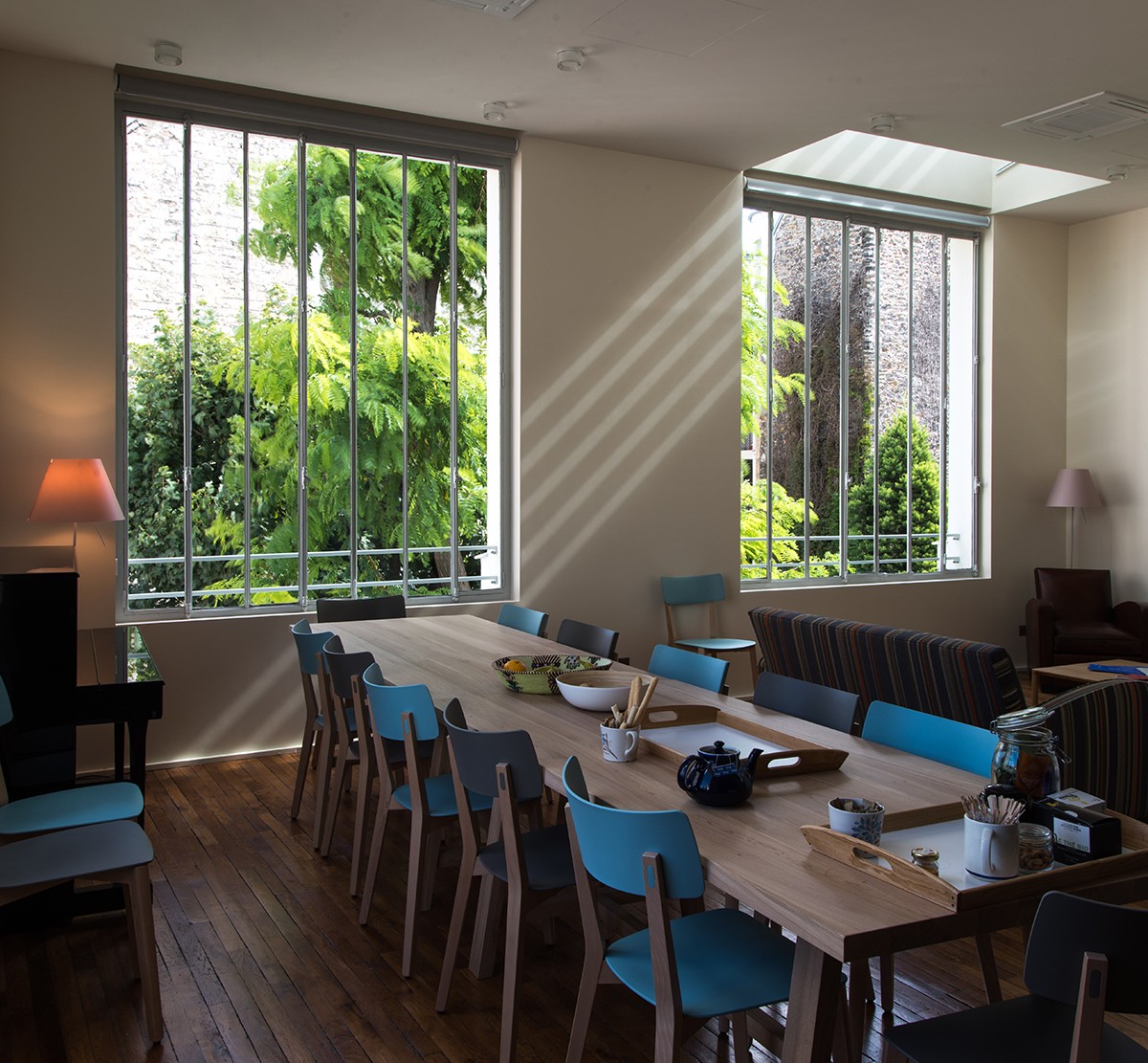 Photo of a room with two big windows with light streaming in; also a long rectangular table surrounded by chairs with a sofa in the background.