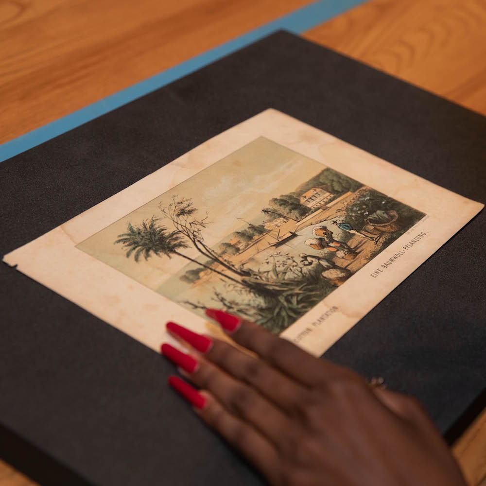 A photo of an old illustration of a town with a pond and a palm tree with a woman's hand holding the image