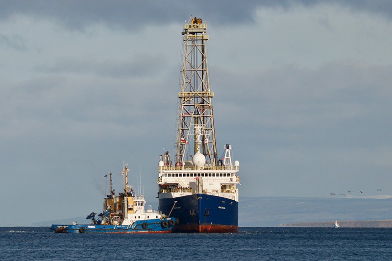RV Joides Resolution in Punta Arenas. Photo by Anieke Brombacher A research vessel with a tower on it is pulled by a tugboat on the ocean.