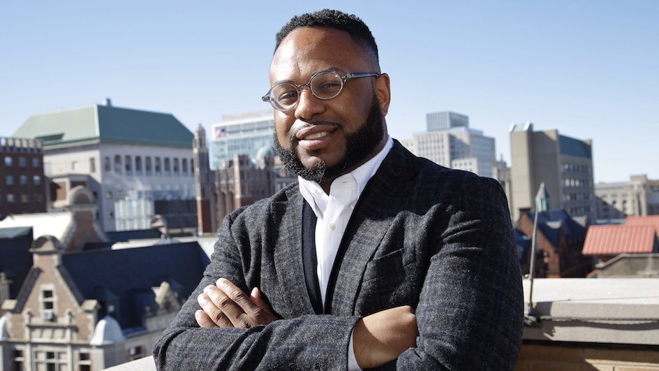 Desmond Patton, associate professor of social work, at Columbia's School of Social Work Desmon Patton in a gray suit, crossing his arms, standing on a rooftop with a view of the tops of other buildings in the background.