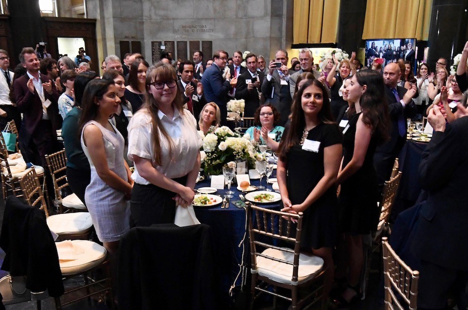 Five young women standing at a table while being applauded by a group of people at a luncheon. 