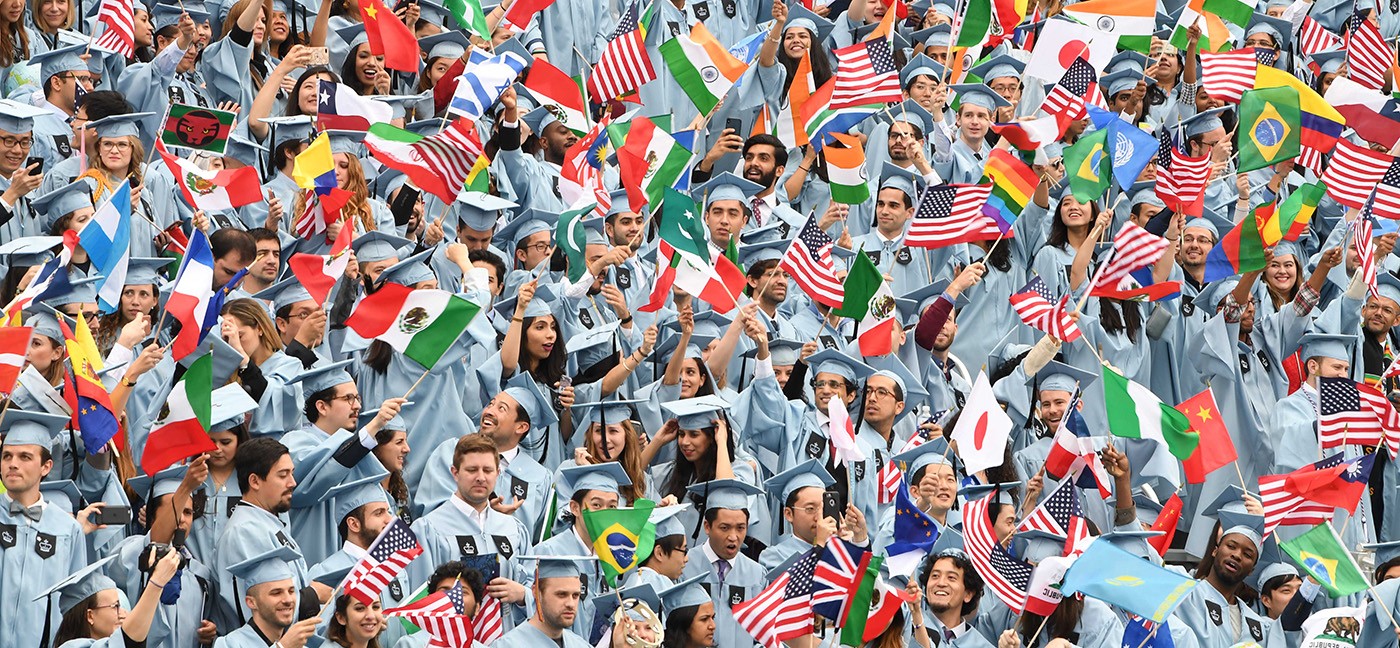 diverse group of students in Columbia blue cap and gowns waving flags from around the world