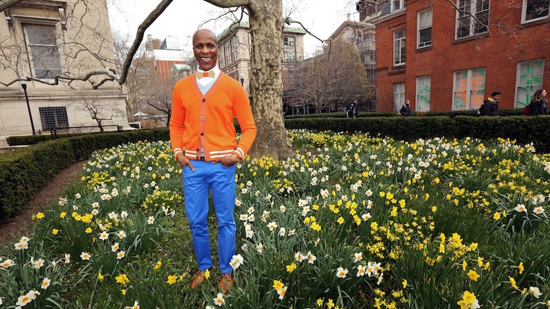 Junio Benjamin in a bright blue pair of pants, an orange sweater, and a yellow bowtie stand in a field of yellow flowers and Columbia buildings in the background. 