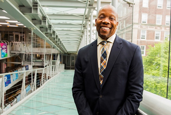 Lance Freeman in a dark gray suit smiling, standing on a glass ramp that looks over a tree on the right and other floors on the left. 