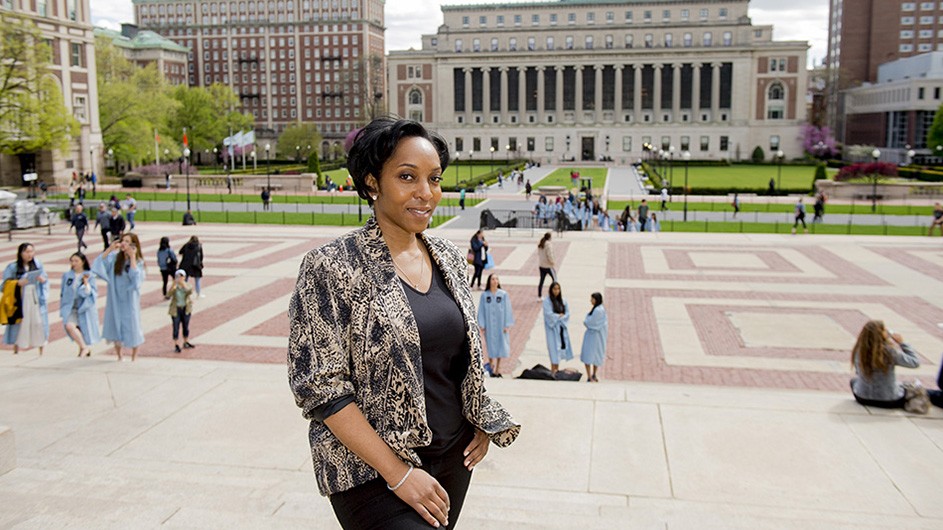 Regan Sommer McCoy, a woman with short dark hair wearing a patterned blouse with a dark T-shirt underneath, faces the camera, with Butler Library, a white stone building with columns, in the background.