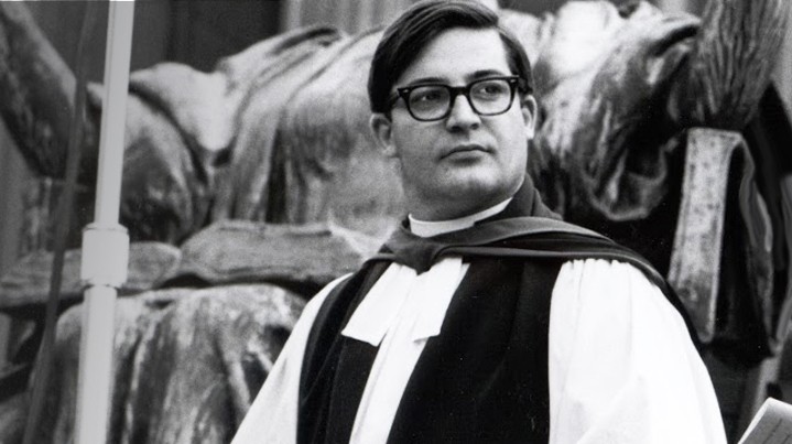 A black and white photo of Chaplain John Dyson Cannon in commencement vestments with an academic hood, standing in front of the Alma Mater statue.