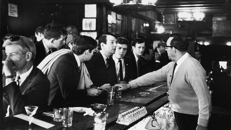 A 1966 image of John Timmons, Dick Leitsch, Craig Rodwell and Randy Wicker sitting at a bar countertop being refused service by a bartender in a white sweater and tie. 
