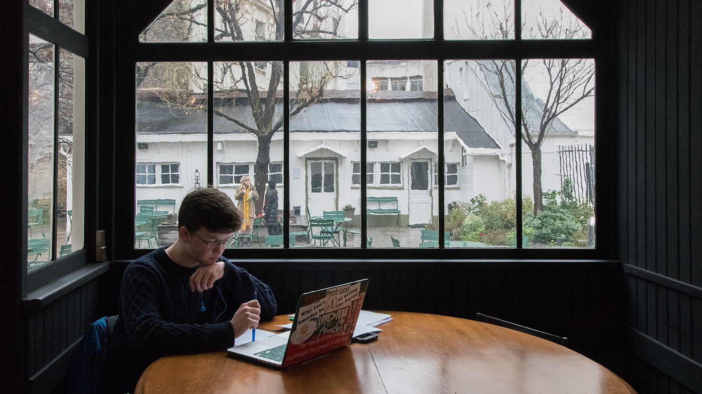 Foyer outside of the Grande Salle, looking out on the second Reid Hall garden (Ferrante Ferranti) Photograph of a young student sitting at a table inside in front of a large window overlooking a garden