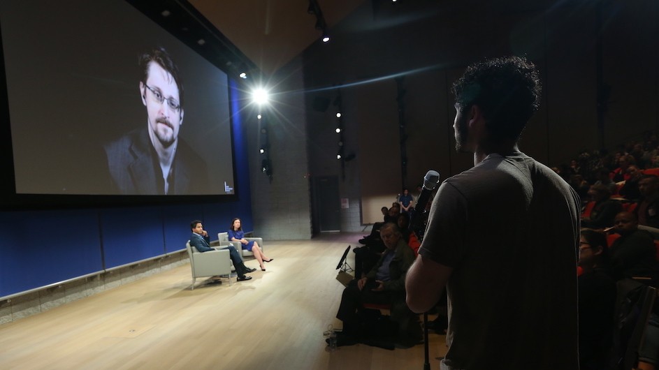 Edward Snowden on a screen in front of two people seated on stage. One person is off to the right on a microphone.