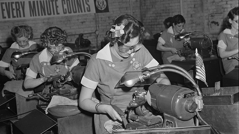 A black and white photo of women in rows sitting at machines putting precision-ground points on drills for a tool company in Chicago during World War II. The women have goggles on and styled hairs. There is a banner on the back wall that says "Every Minute Counts.”