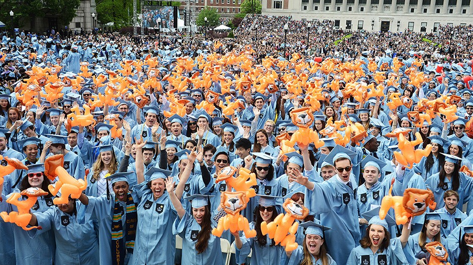 A diverse group of students in their Columbia blue caps and gowns celebrating commencement while holding up inflatable Roaree the Lion mascots.