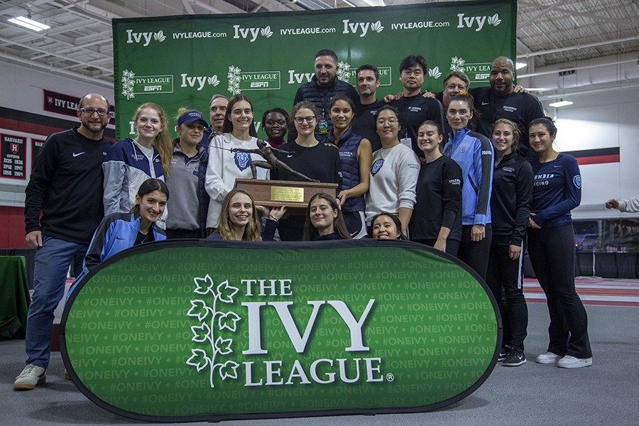 22 fencing athletes and coaches pose with a championship trophy against a green Ivy League athletics backdrop in a fencing room.