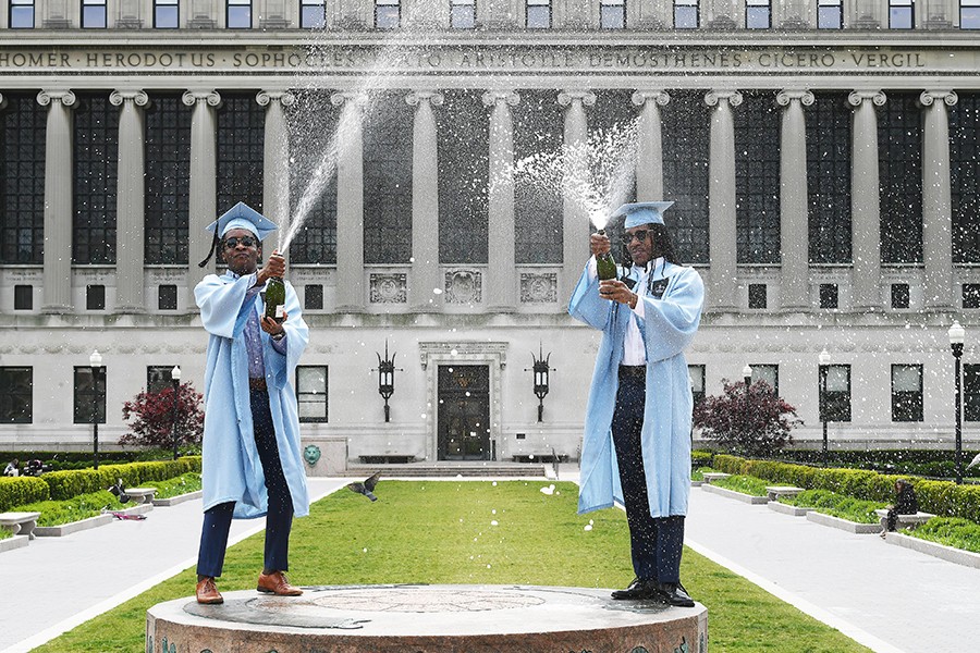 Two Columbia graduates in graduation caps and gowns pop champagne in front of Butler Library. 