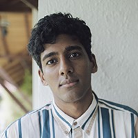 headshot of Anuk Arudpragasam, medium skinned man with hair in a blue and white striped collared shirt