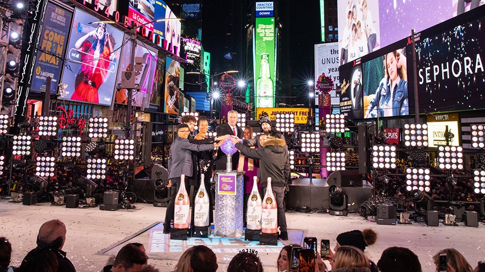 Jared Fox (wearing a hat), BioBus board member and Teachers College alumnus, drops the Times Square ball with two of his students, Mayor Bill de Blasio (center) and First Lady Chirlane McCray (to the right of the mayor), as well as fellow teacher Aida Rosenbaum (left of the mayor) and two of her students. Photo by Amy Hart, Courtesy of Times Square Alliance NYC Mayor Bill de Blasio and First Lady Chirlane McCray stand on a platform in the middle of Times Square with teachers Jared Fox and Aida Rosenbaum accompanied by two students each.