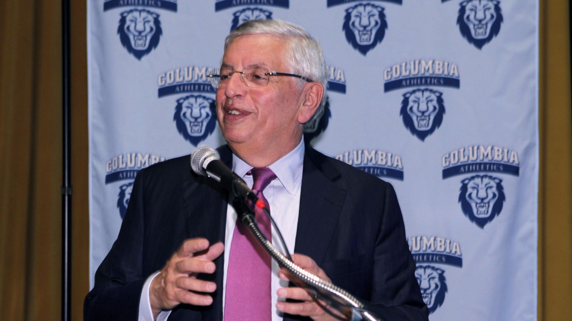 Photo of David Stern in a navy blue suit, behind a microphone, in front of a poster that repeats Columbia Athletics