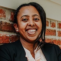 dark-skinned woman with dark hair, in a dark suit and light peach shirt.