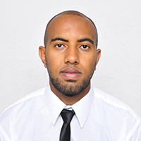 Headshot of a dark-skinned man with short, dark hair and dark facial hair, in a white shirt and dark die