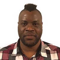 Dark-skinned man with a dark mohawk, dark beard in a red, cream, and brown checked shirt.