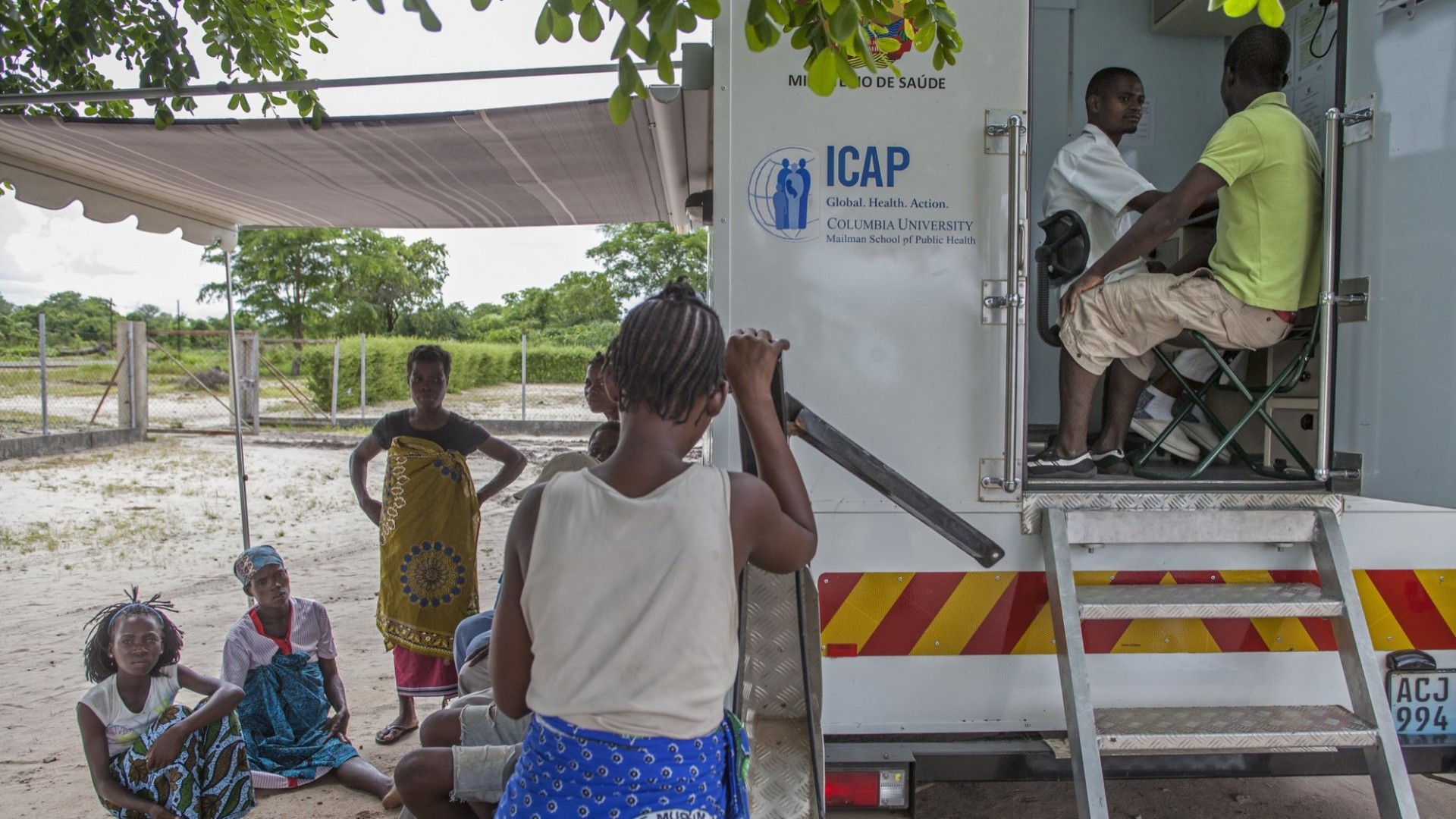 Residents in Mozambique receive health services at a mobile clinic supported by ICAP at Columbia University. The clinic, funded by the President’s Emergency Fund for AIDS Relief, provides HIV testing and care to the community. Patients sitting on dirt ground or standing by a white truck with an open awning on the side and a door at the back revealing a medical worker talking to a patient.