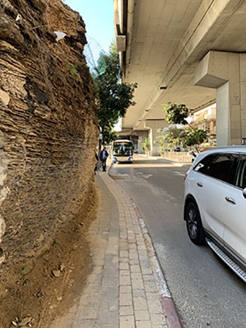 A cliff sticks out from a sidewalk and street that is under a elevated roadway.