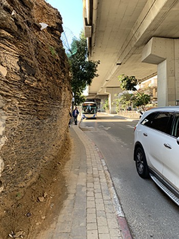 Nature and urban design, cheek by jowl, in Tel Aviv. A cliff sticks out from a sidewalk and street that is under a elevated roadway.